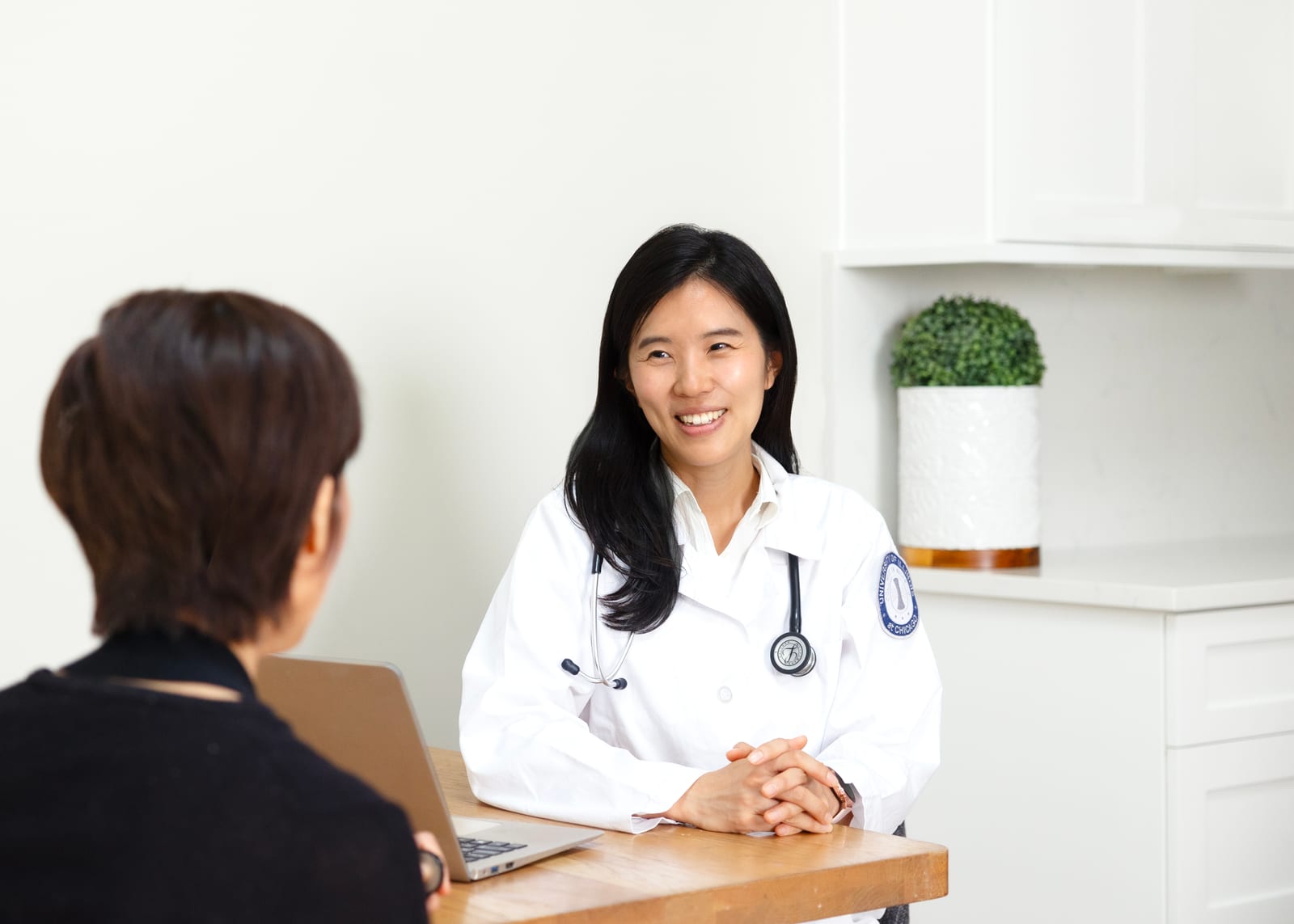 Physician in a white coat with a stethoscope, leaning in to listen to a patient across a bright consulting desk