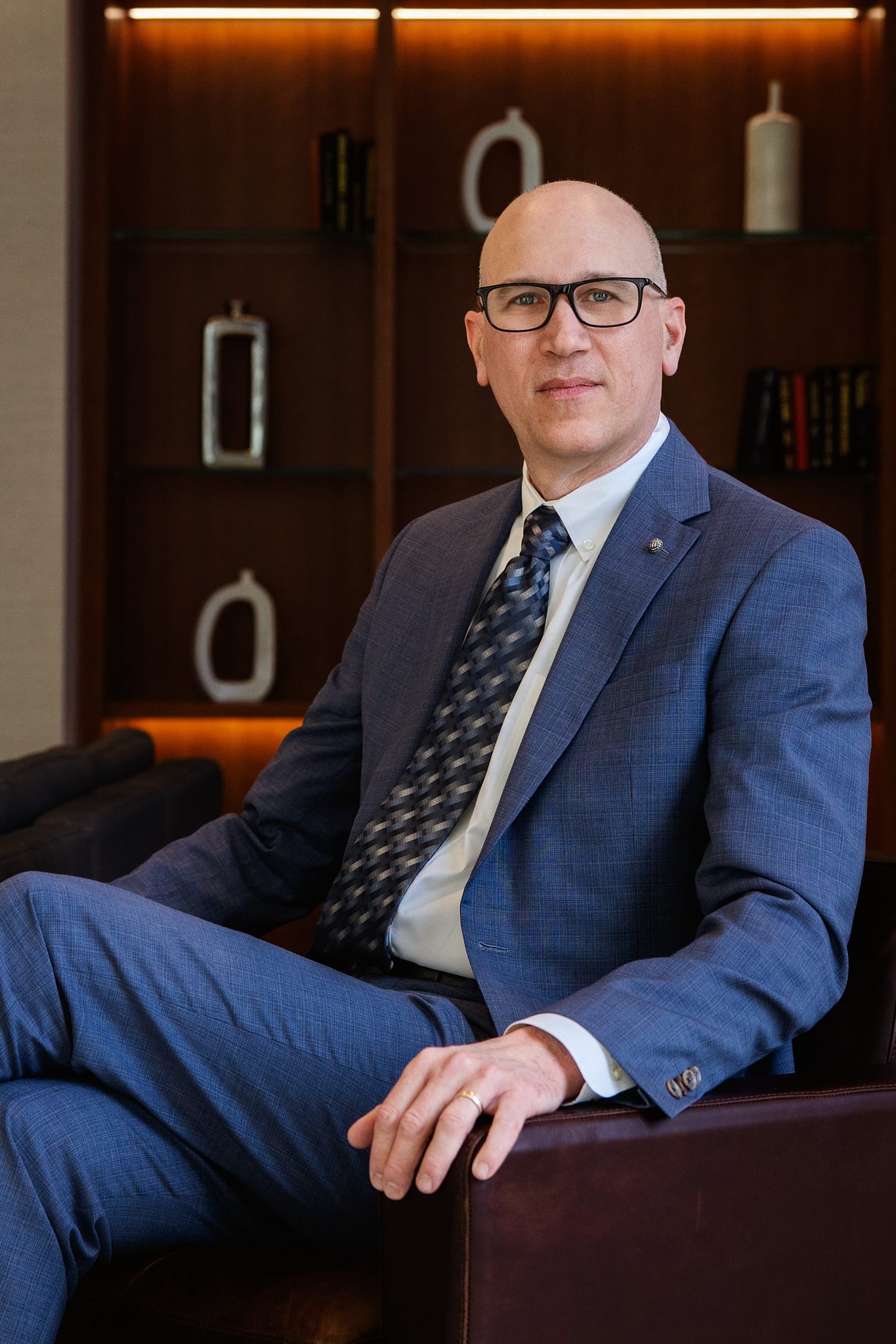 Executive in a blue check suit seated on a leather sofa in front of a warmly lit walnut bookcase