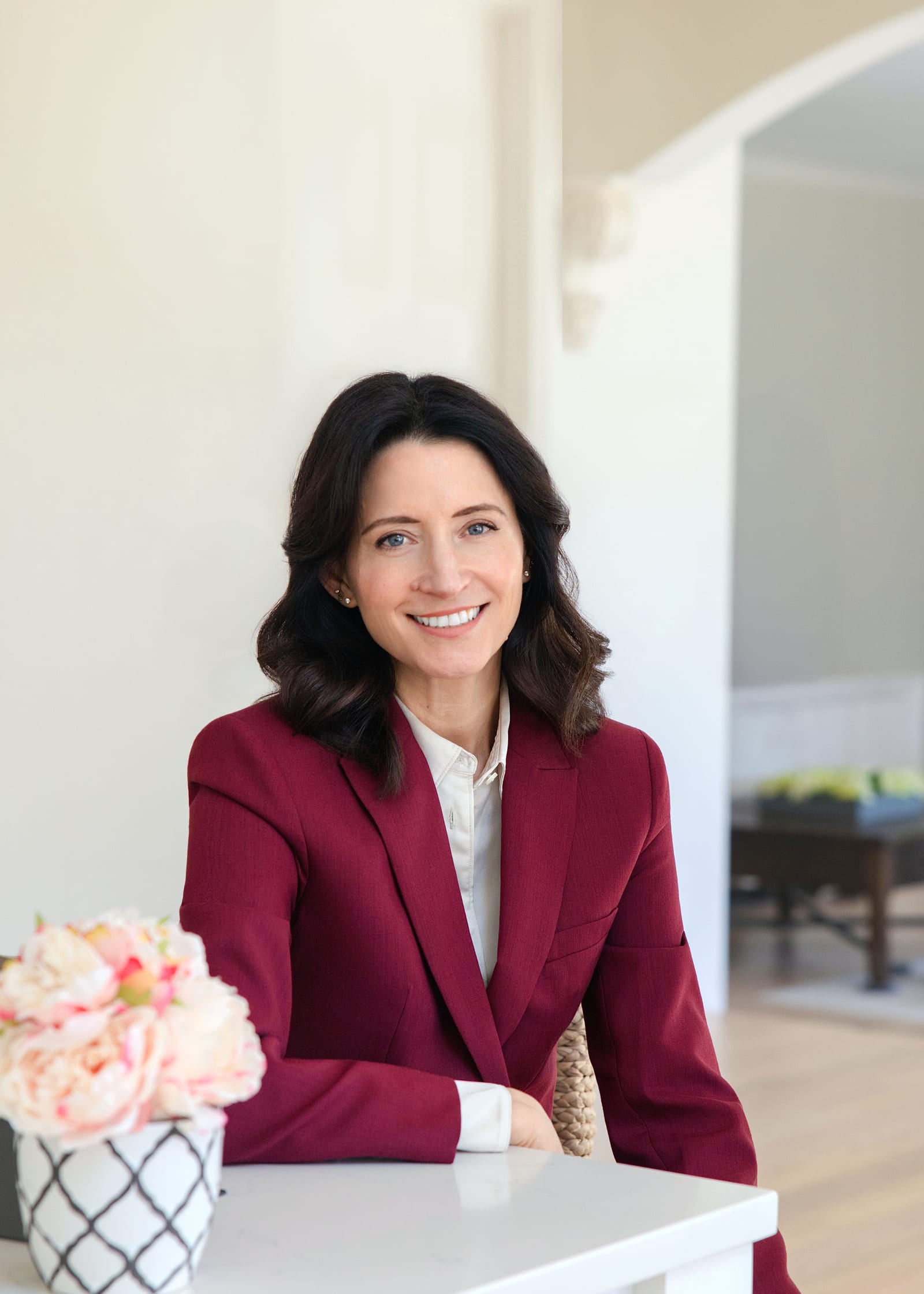 Founder in a burgundy blazer seated at a white table beside a vase of peonies