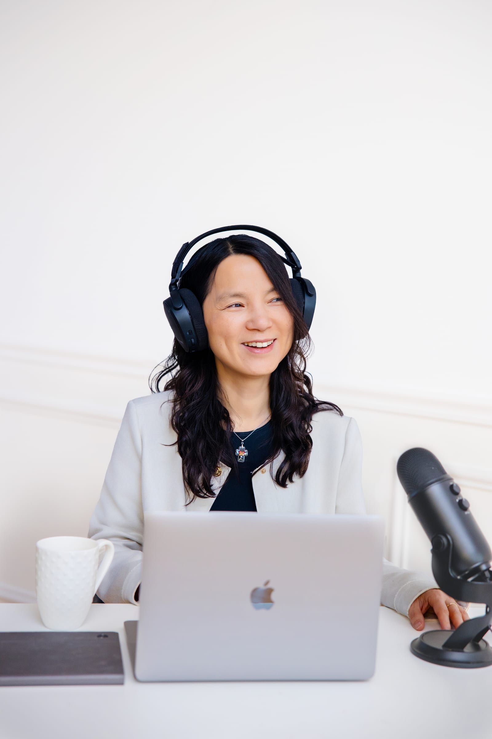 Podcaster in headphones and a white blazer laughing at a bright desk with a microphone and laptop