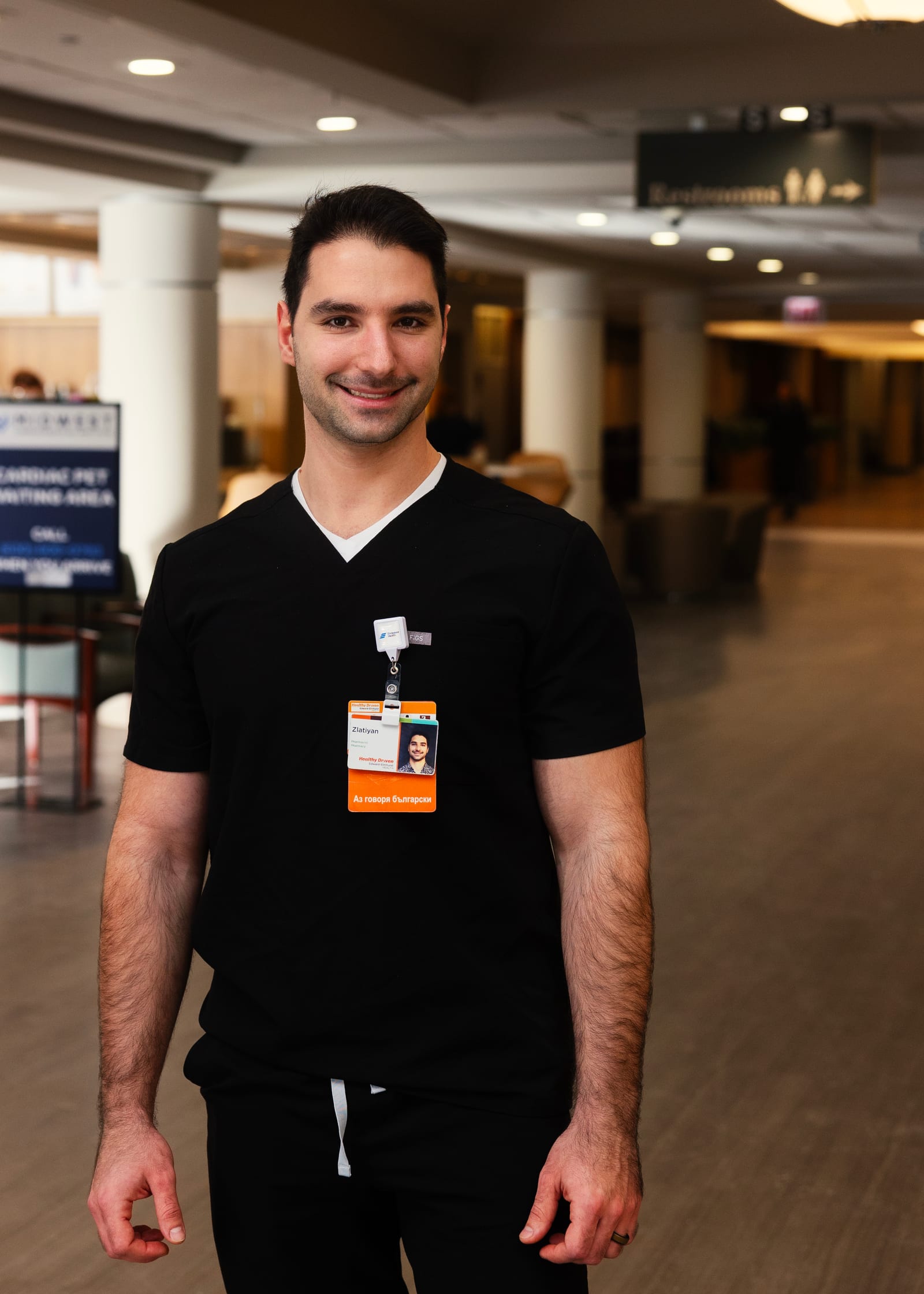 Clinician in black scrubs with a hospital badge smiling in a bright hospital lobby