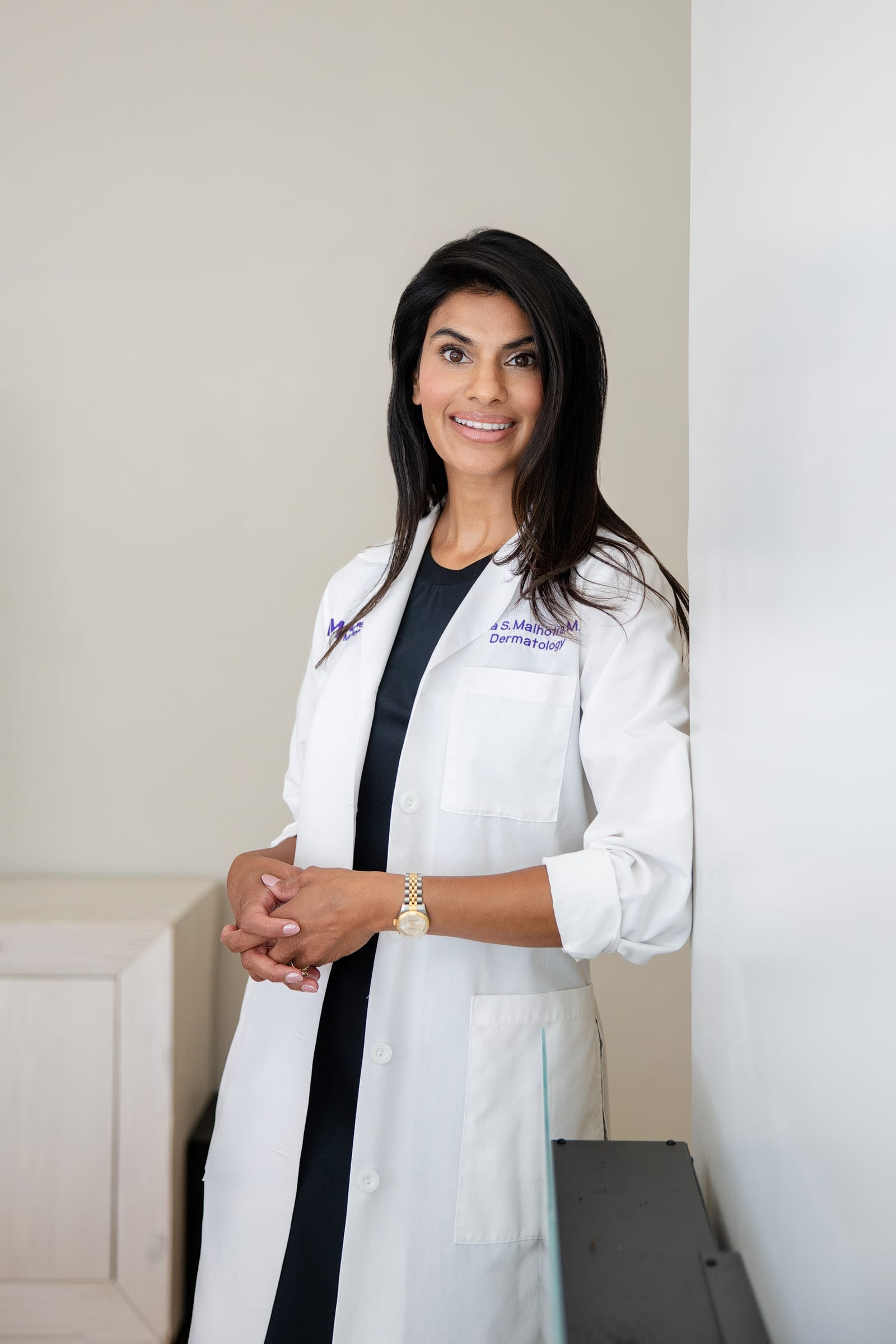 Clinician in a white coat over black, hands clasped, standing against a soft grey wall