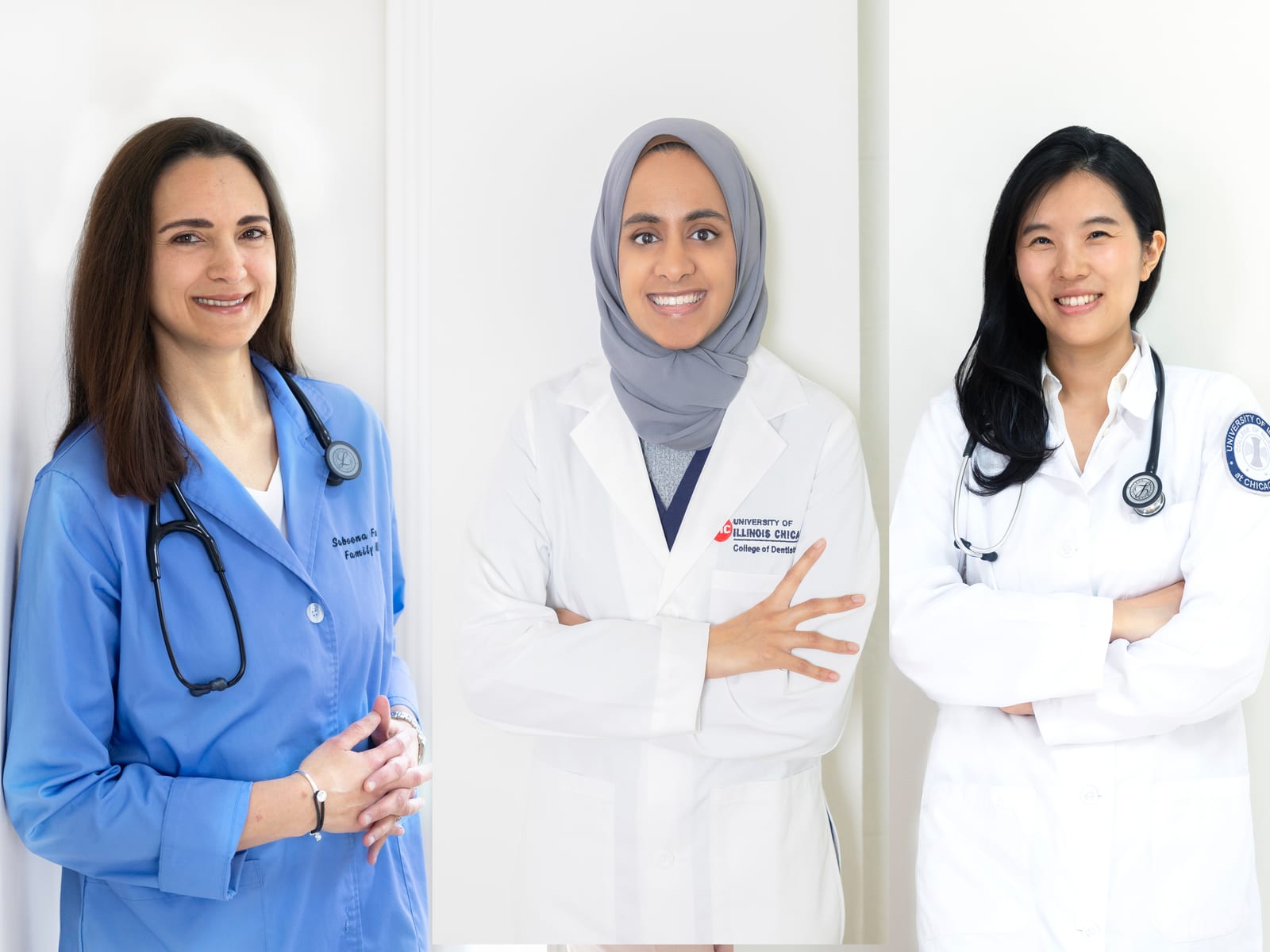 Three women clinicians in white coats and blue scrubs with stethoscopes smiling against a white wall