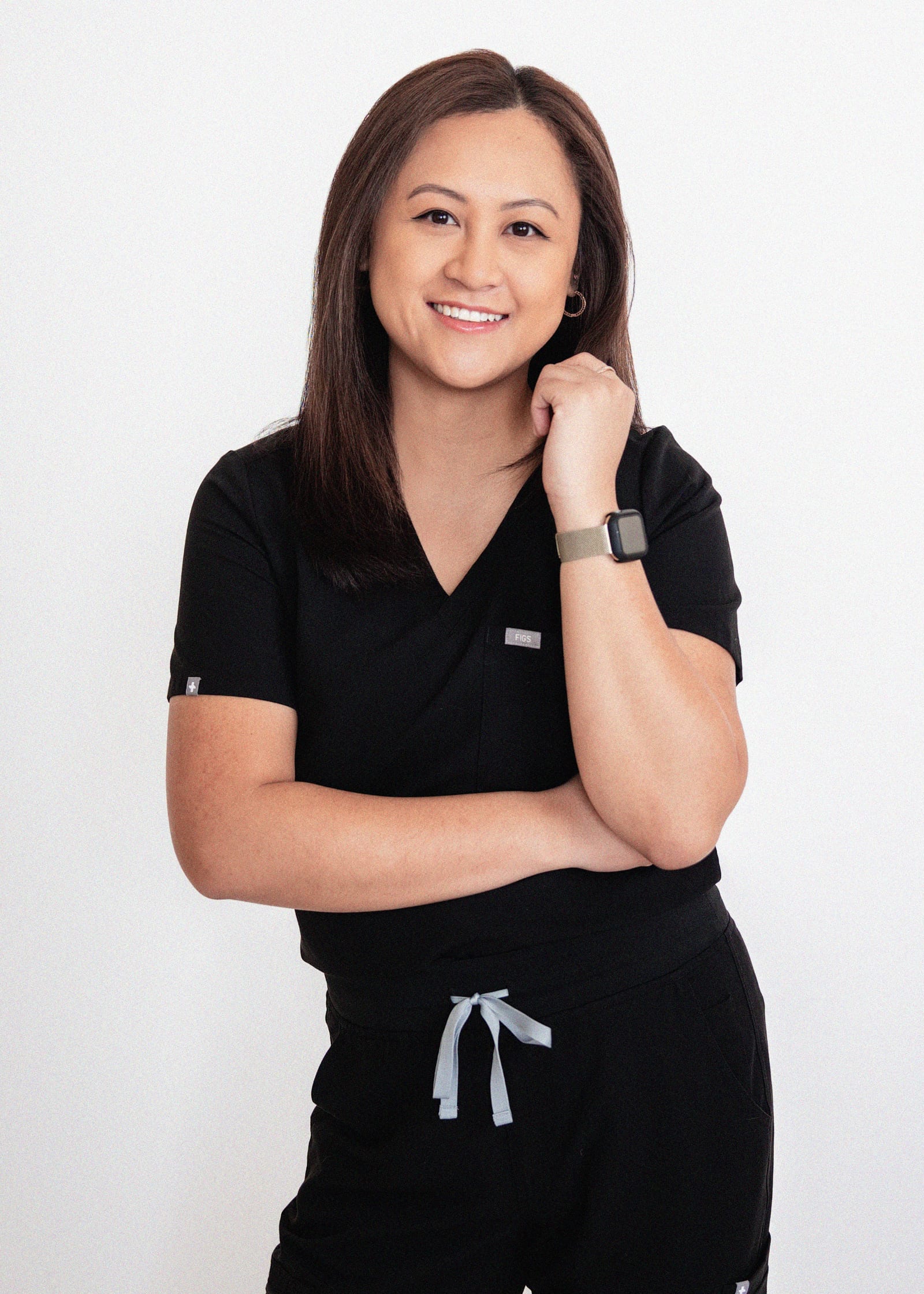 Clinician in black scrubs smiling with one arm folded and a hand raised to her chin, against a plain white wall