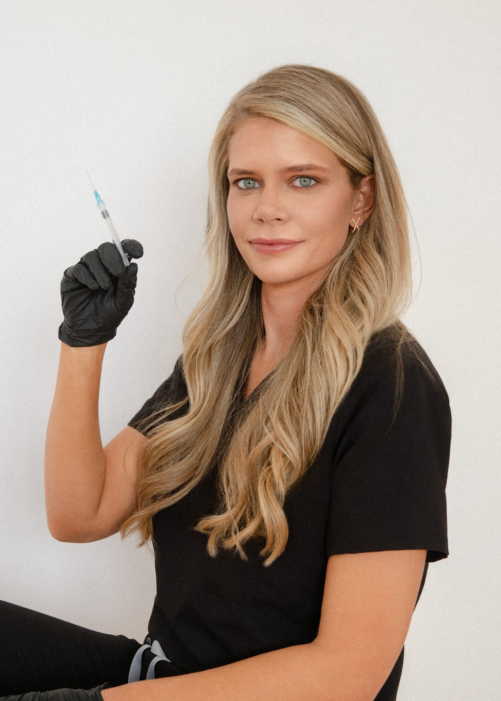 Aesthetic practitioner in black scrubs and gloves holding a syringe, against a white wall