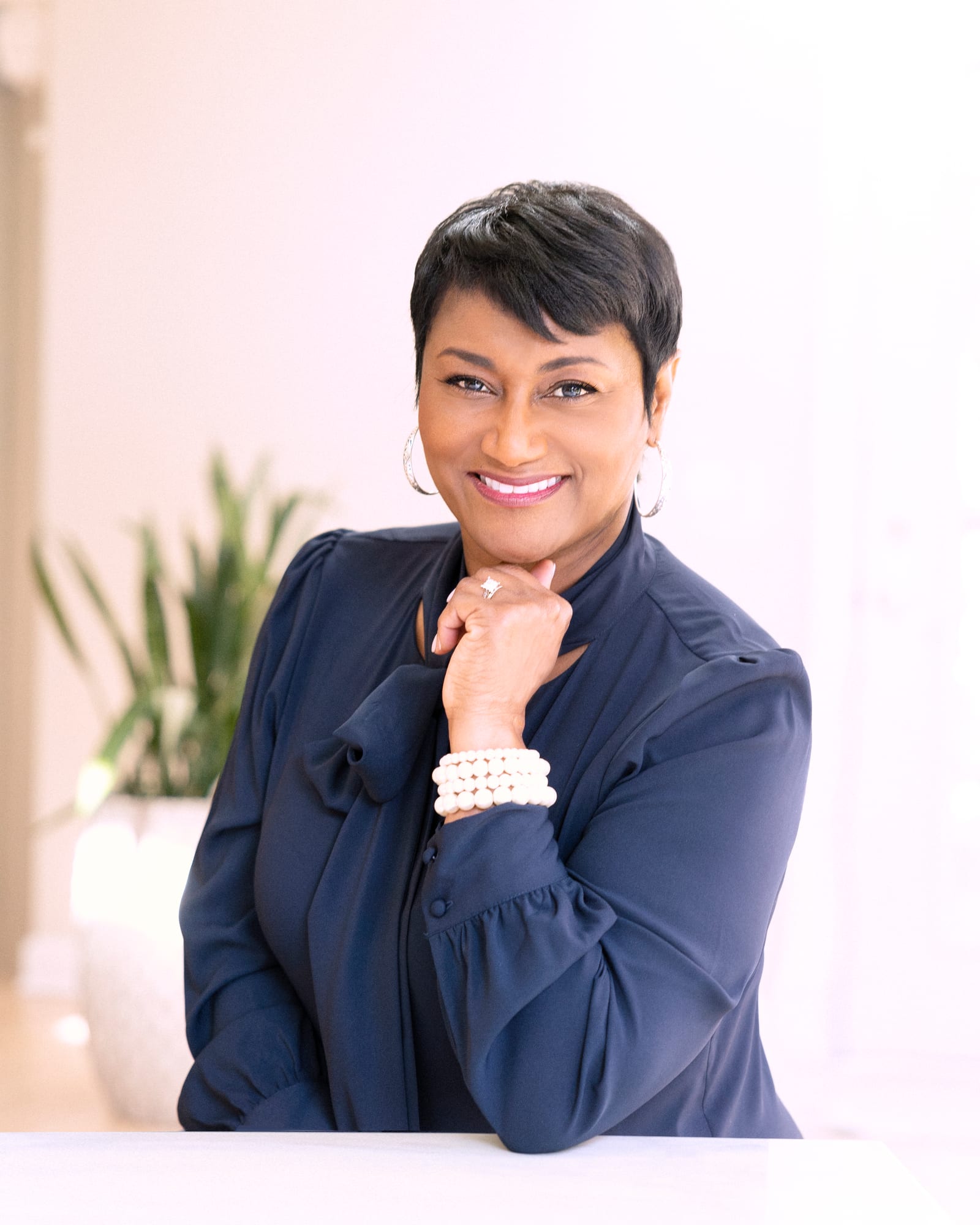 Executive in a navy blouse with pearl bracelet, chin on hand, smiling in a bright, airy room