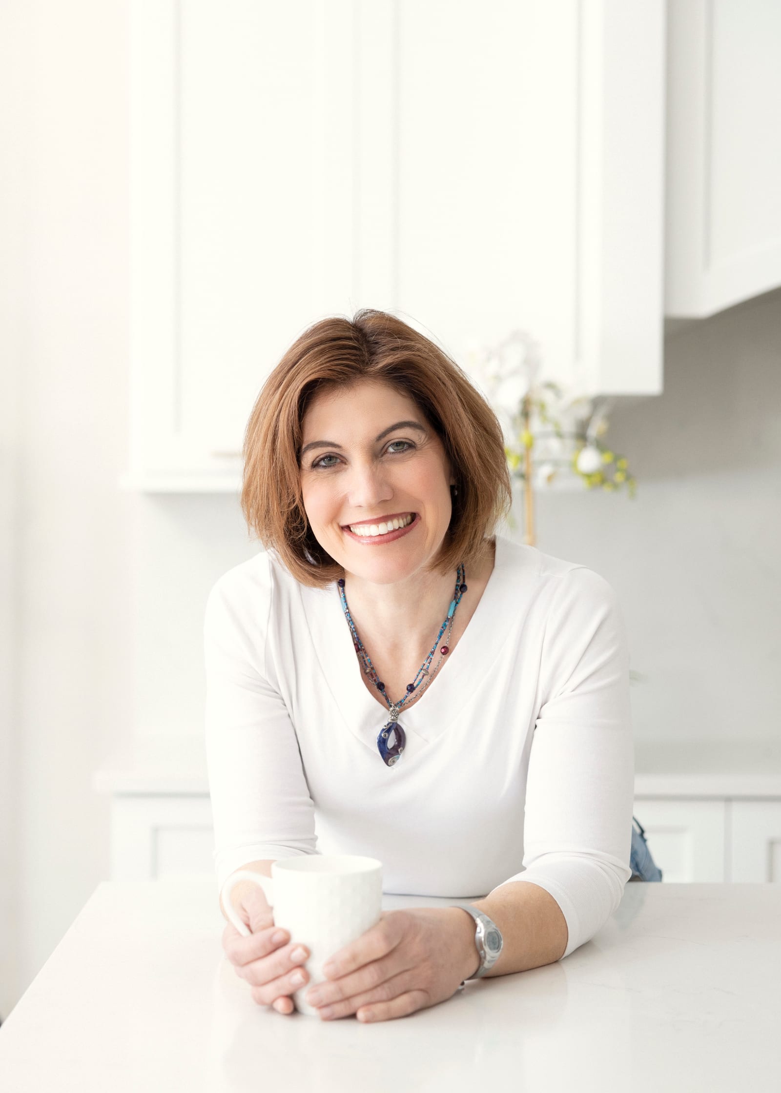 Smiling business owner in a white top and beaded necklace cradling a mug at the counter of a sunlit white kitchen