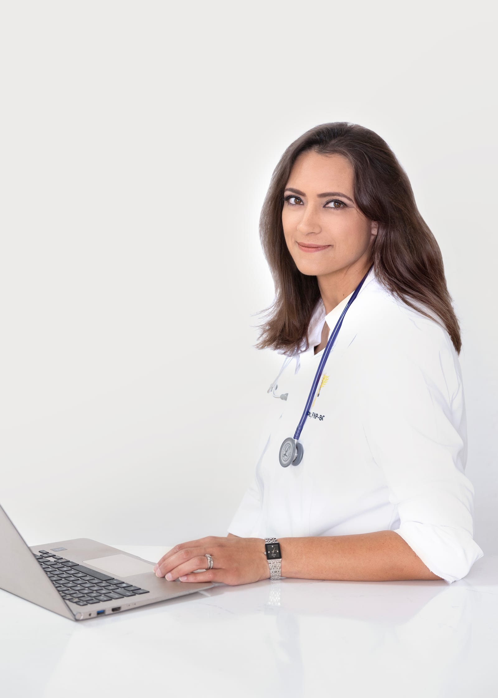 Nurse practitioner in a white coat with a purple stethoscope turning from her laptop with a soft smile, high-key white set