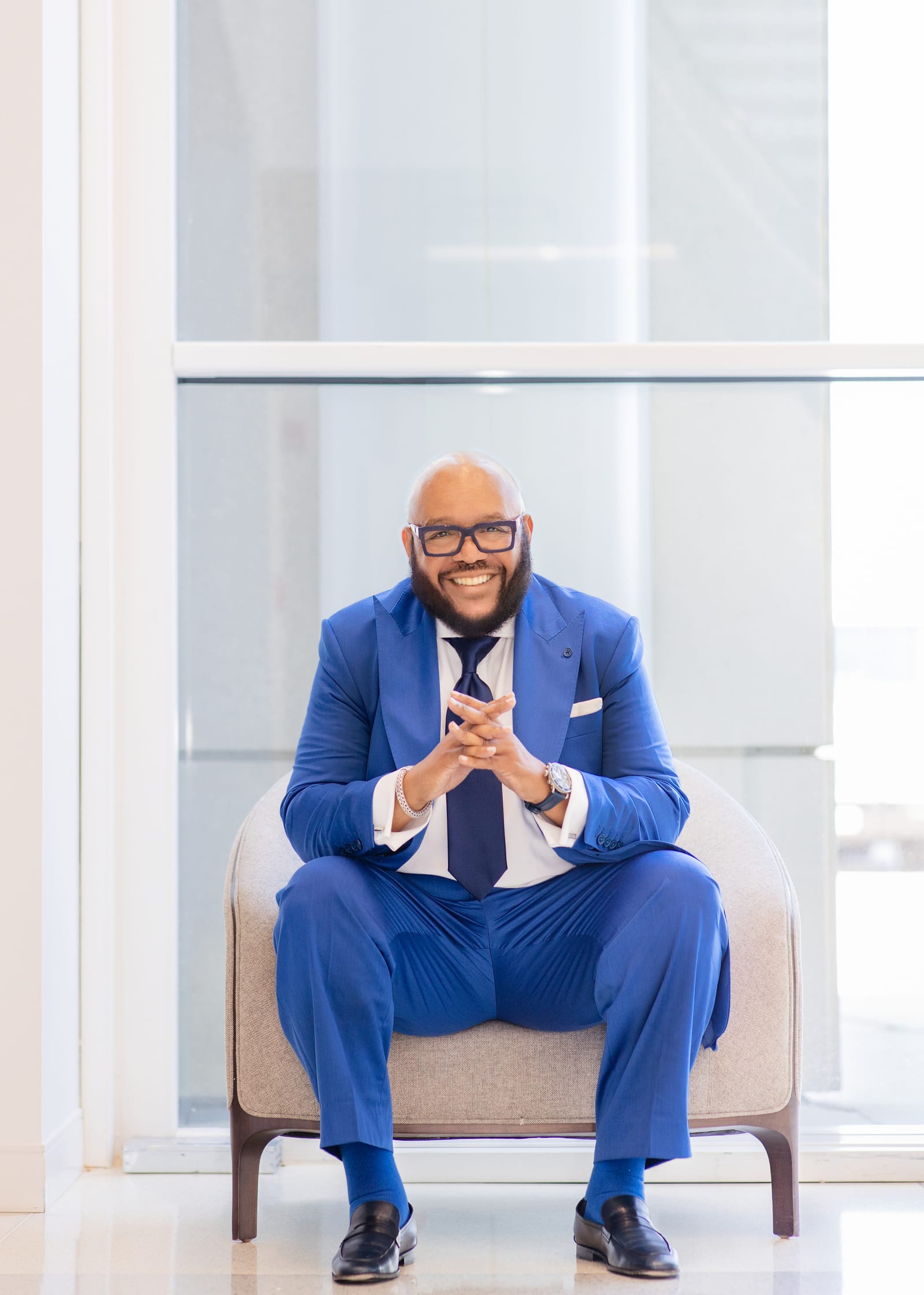 Executive in a bright blue suit and navy tie seated in an armchair against a wall of frosted glass