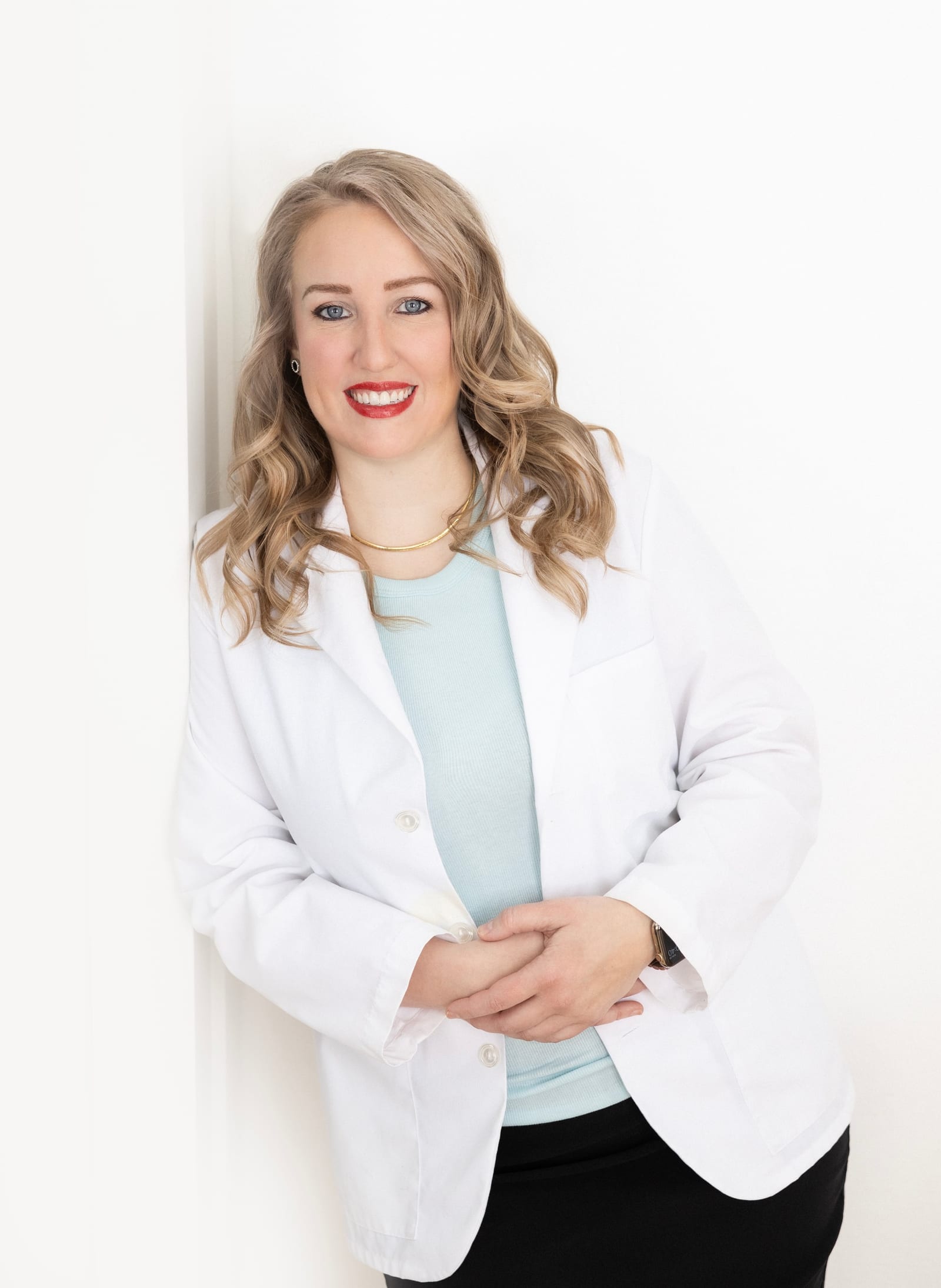 Clinician in a white coat over a mint top, hands clasped, leaning against a white wall with a broad smile
