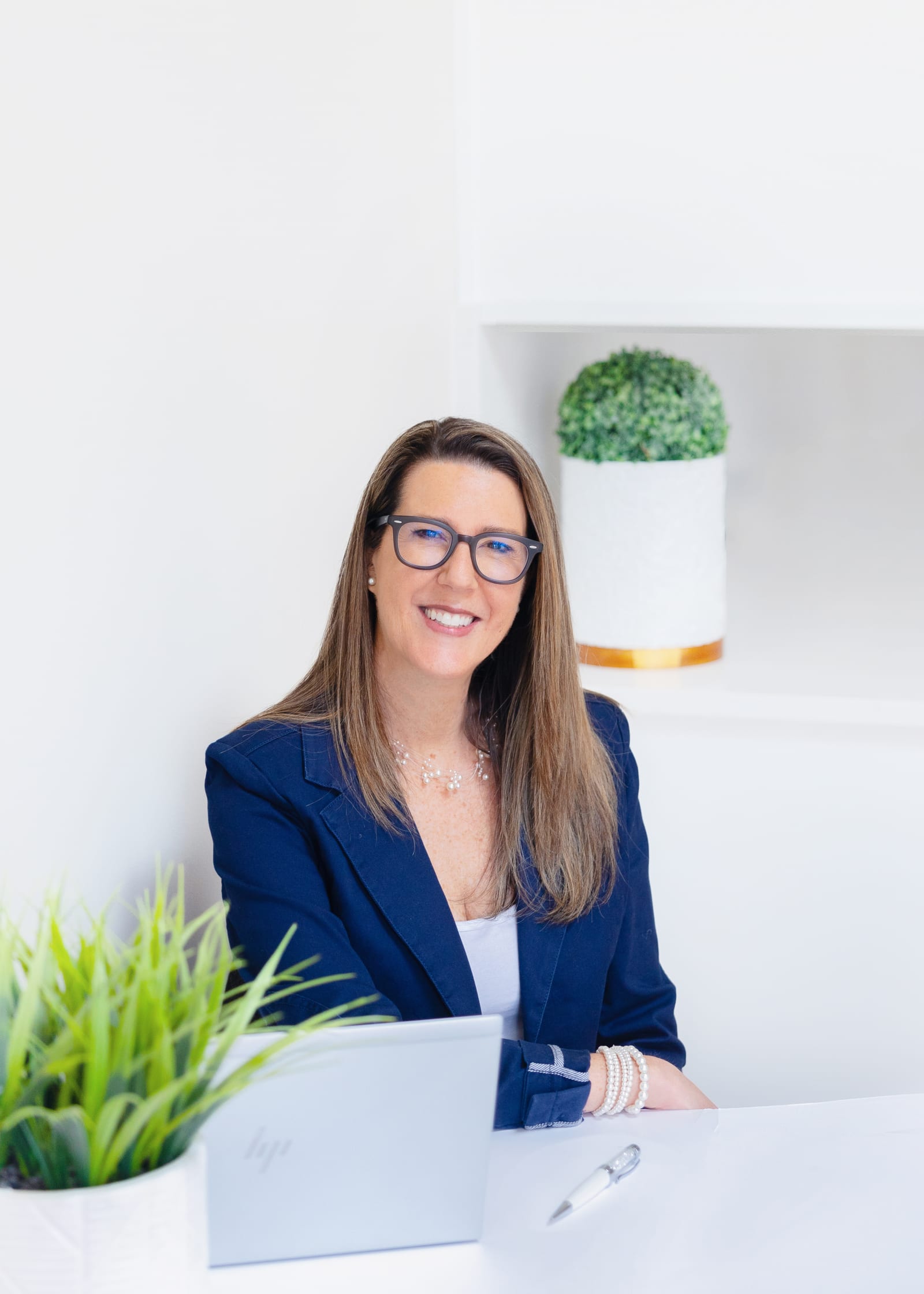 Business owner in a navy blazer and glasses seated at a bright white desk with a laptop and a plant