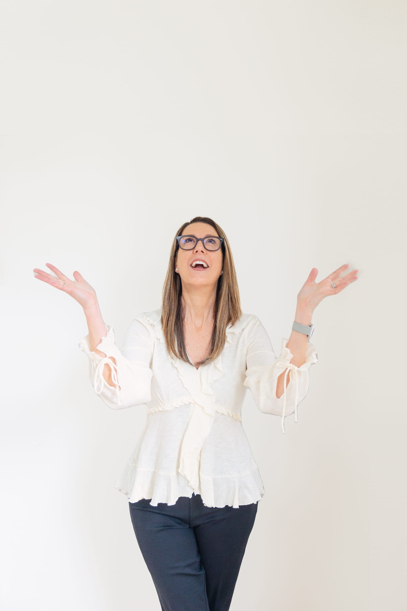 Business owner in a cream ruffled top laughing with both arms raised, looking up, against a plain white wall