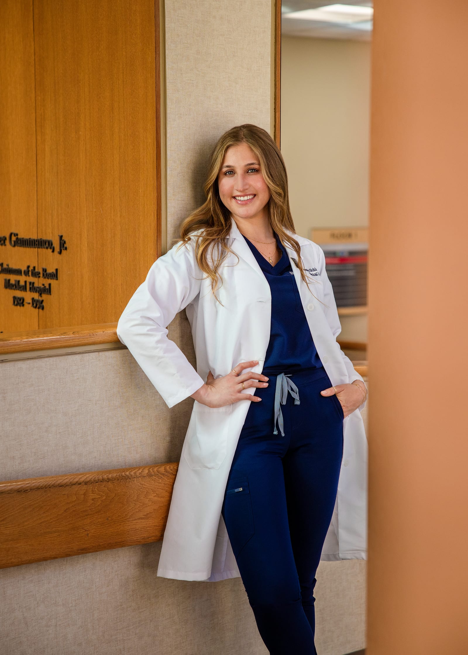 Clinician in a white coat over navy scrubs, hand on hip, leaning against a panelled corridor wall