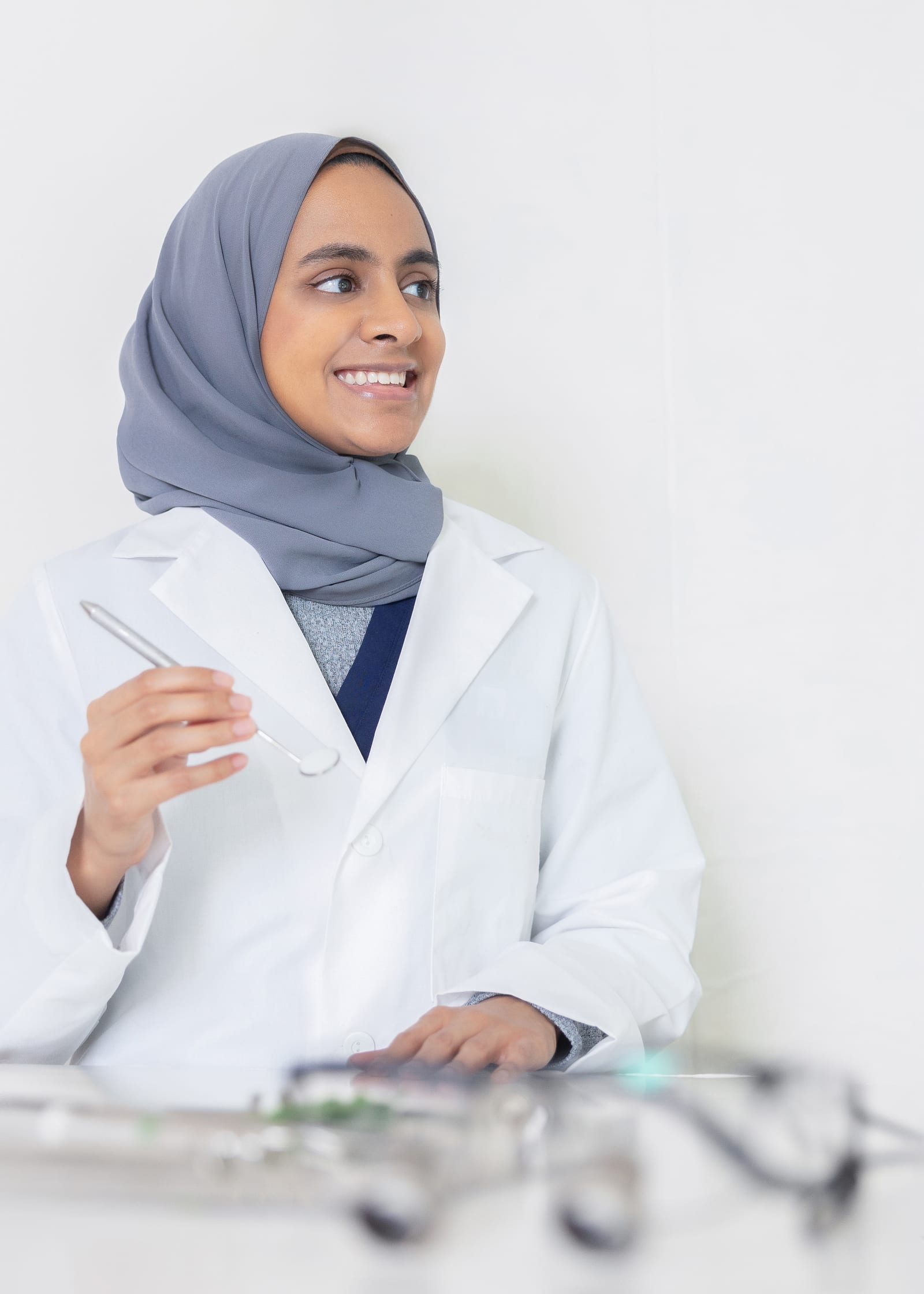 Dentist in a grey hijab and white coat holding a dental mirror at a bright treatment desk