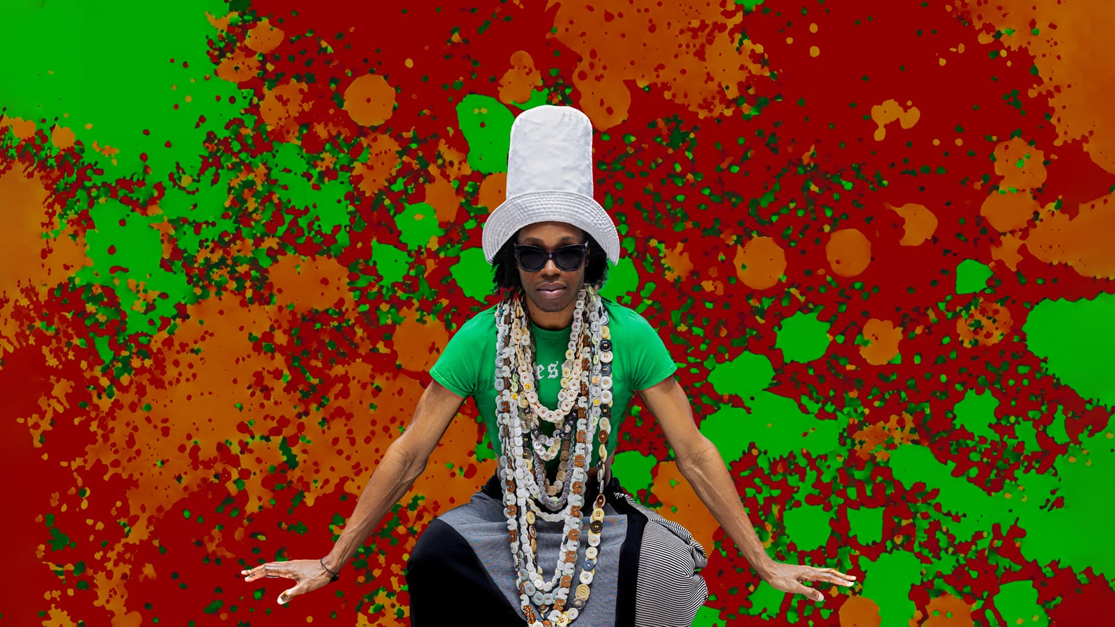 Artist in a white bucket hat, sunglasses and layered bead necklaces, arms open before a red and green paint-splattered backdrop