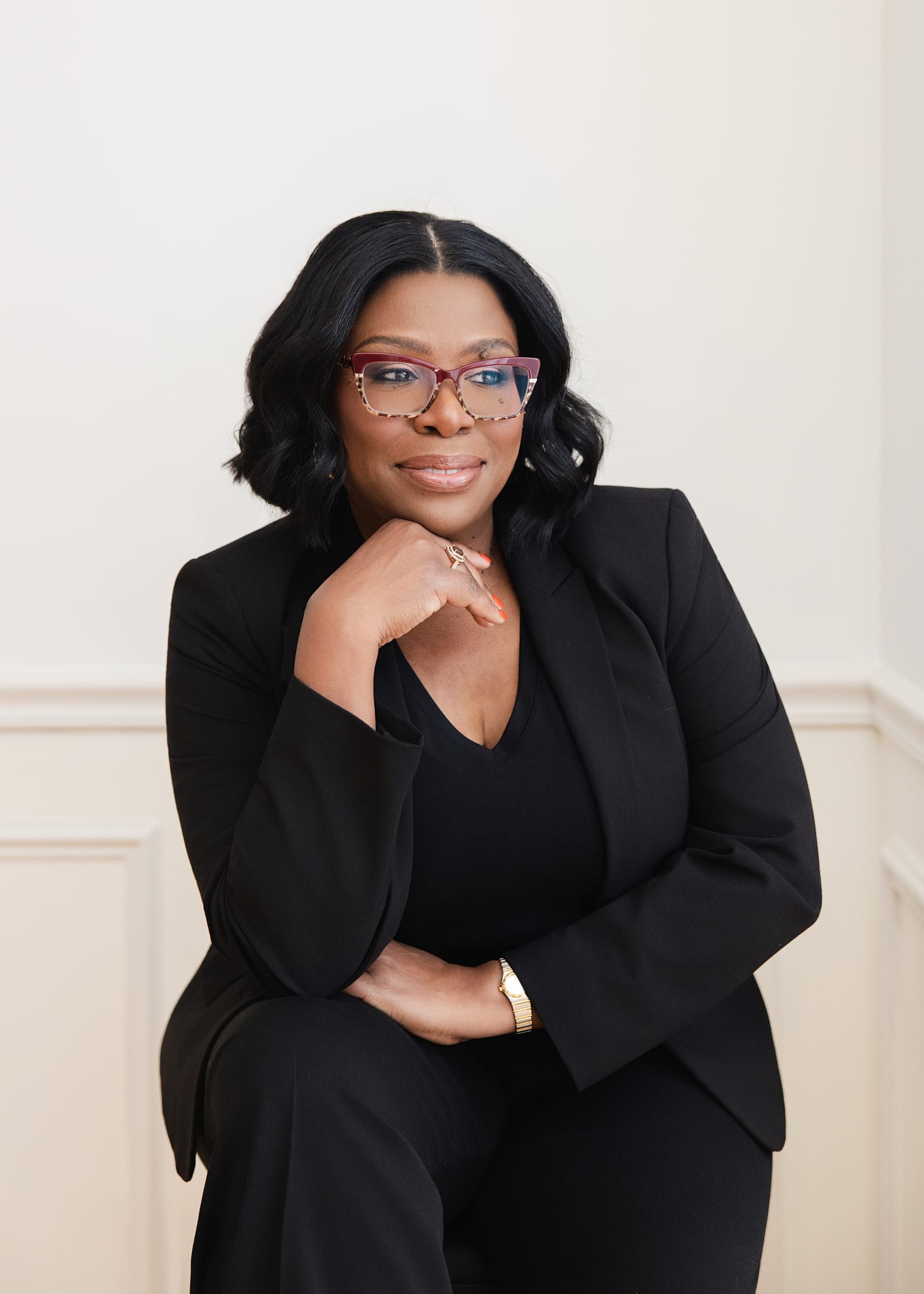 Professional in a black suit and glasses, chin resting on her hand, against a white wall