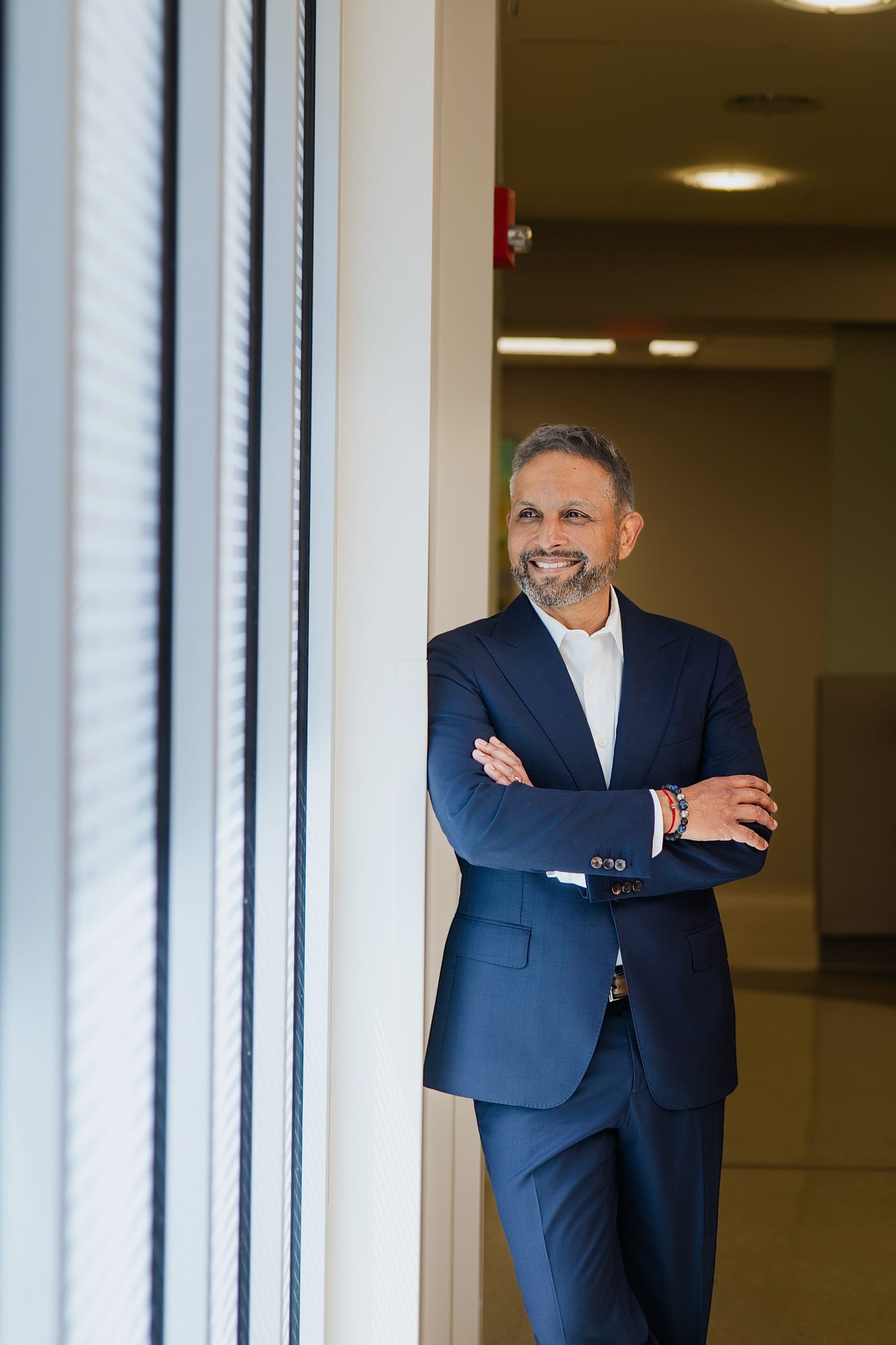 Executive in a navy suit with arms crossed, leaning in a bright doorway beside a run of window blinds