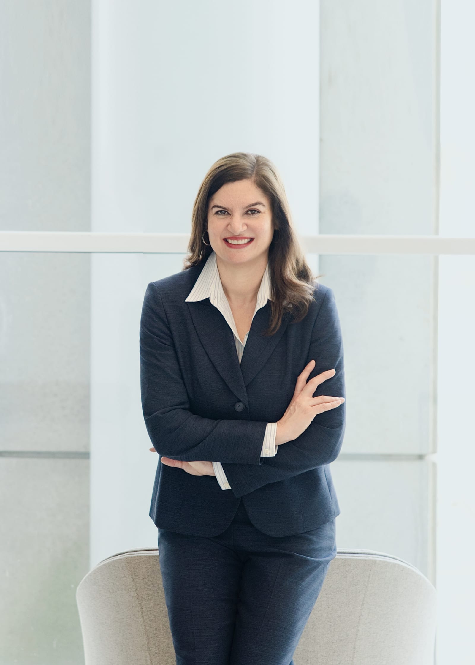 Executive in a charcoal suit and pinstripe shirt, arms crossed, standing against a pale glass wall