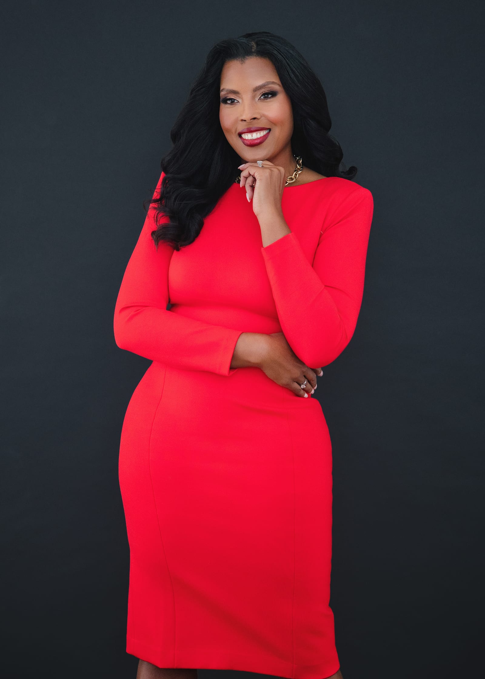 Executive in a scarlet dress, hand to her chin, against a near-black ground