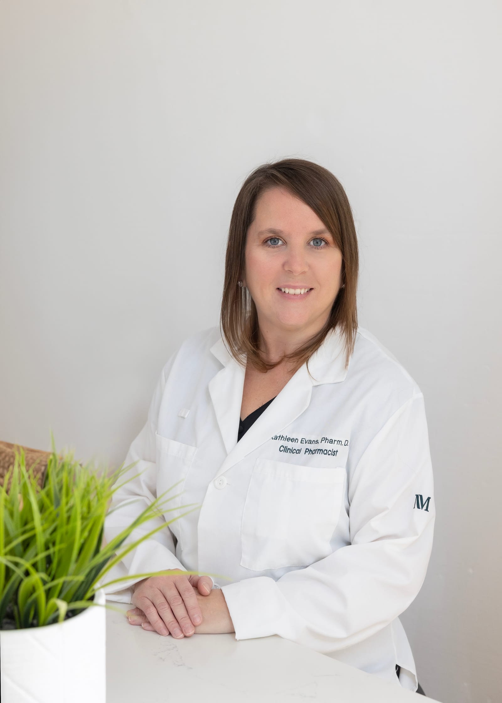 Clinical pharmacist in a white coat standing beside a plant against a white wall