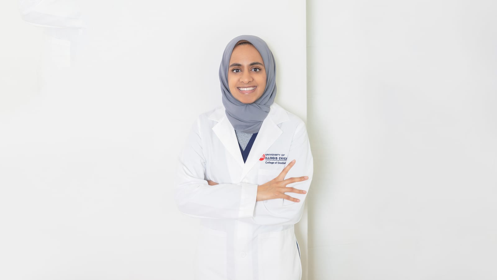 Physician wearing a hijab and white coat, arms folded, standing against a white wall