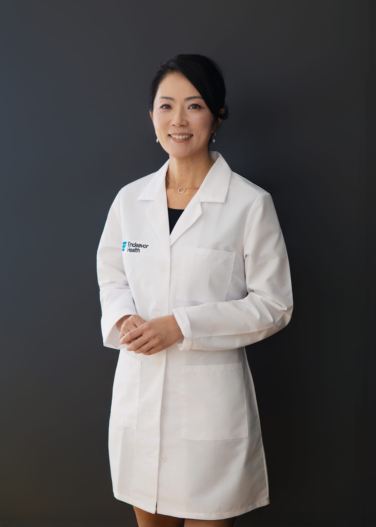 Clinician in an Endeavor Health white coat standing with hands lightly clasped against a dark grey ground