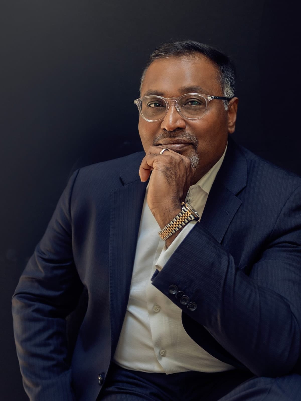 Executive in a navy pinstripe suit and clear-framed glasses, hand to chin, against a near-black ground