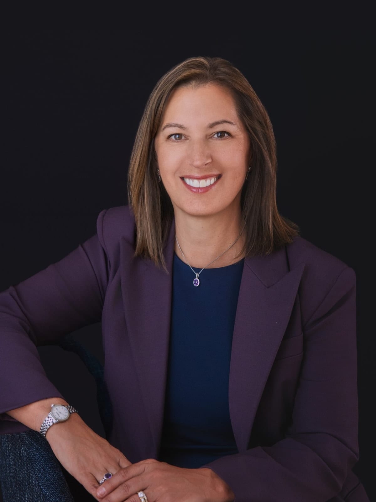 Executive in a plum blazer over a navy dress, seated, against a near-black ground