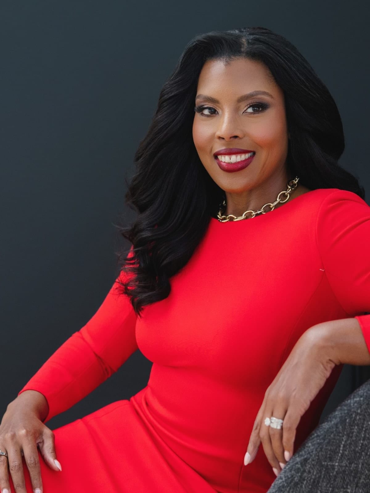 Executive in a red dress and gold chain, seated and turned to camera, against a charcoal ground