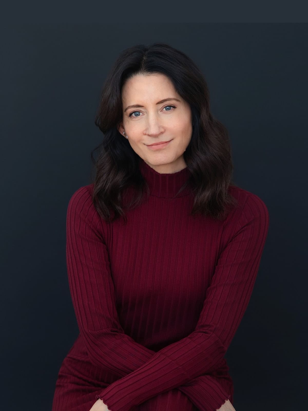 Executive in a burgundy ribbed turtleneck, seated with arms folded, against a deep navy ground