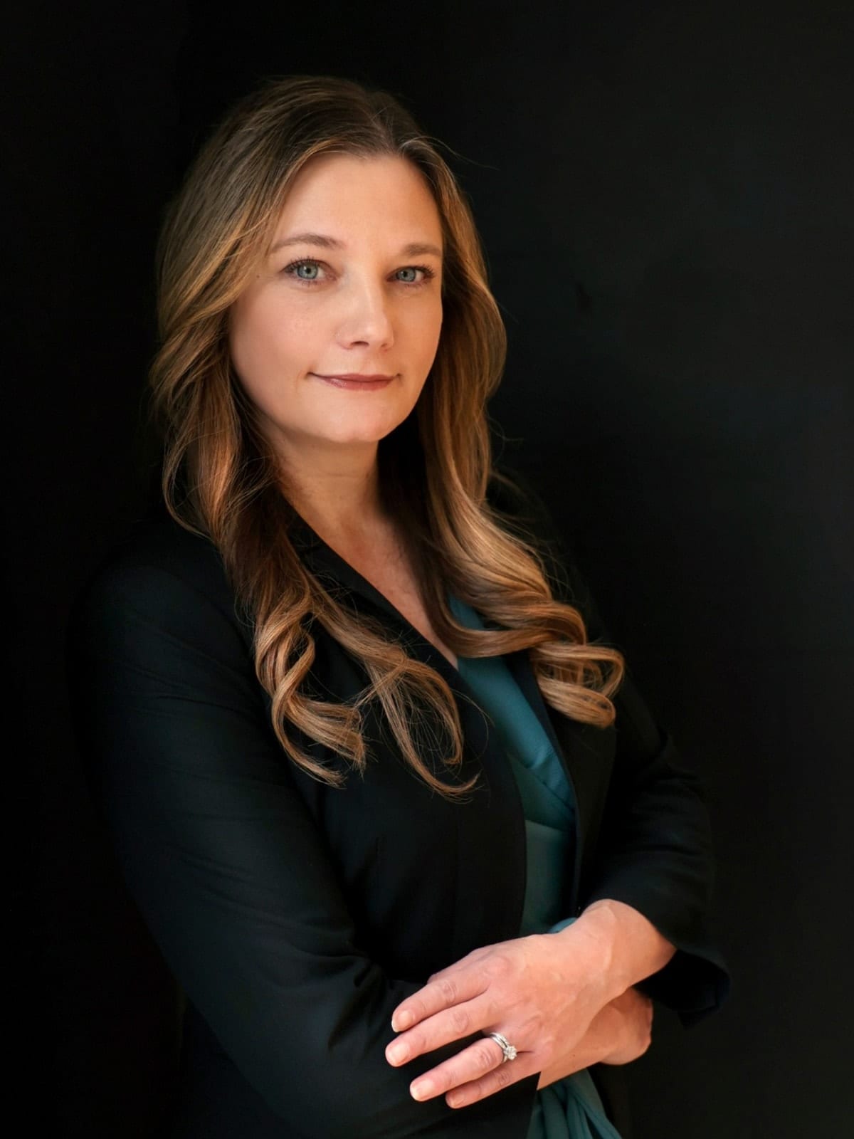 Executive in a black suit over a teal blouse, arms folded, against a black ground