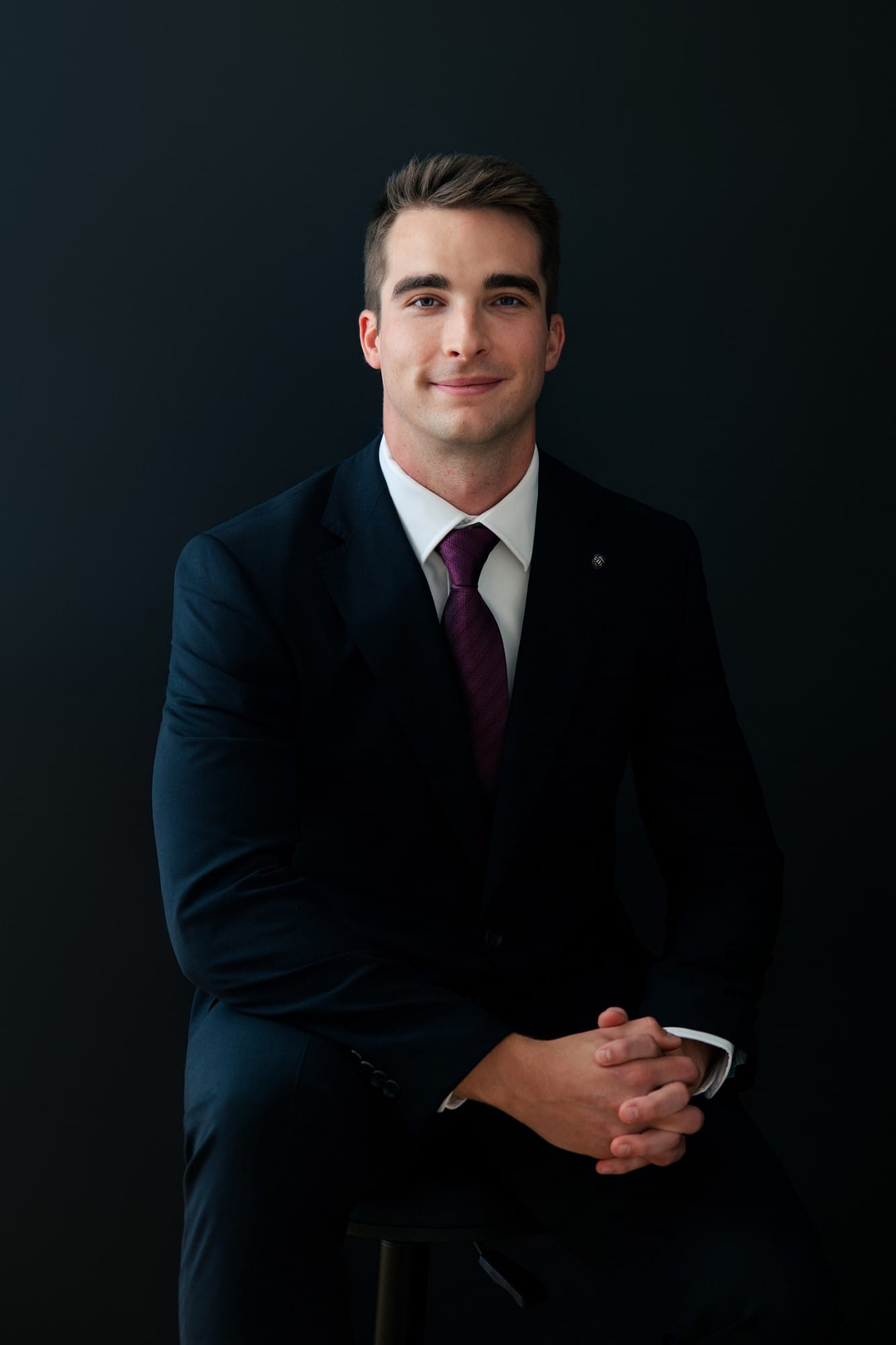 Professional in a navy suit and burgundy tie, seated with hands clasped, against a near-black ground