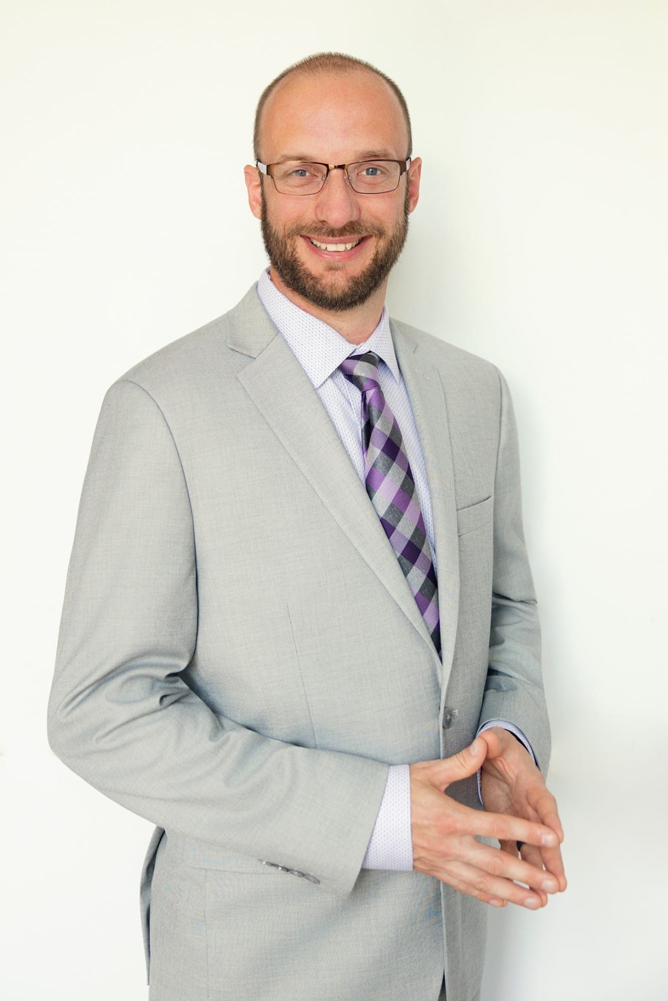 Professional in a light grey suit and checked tie, glasses, hands together, against a white ground