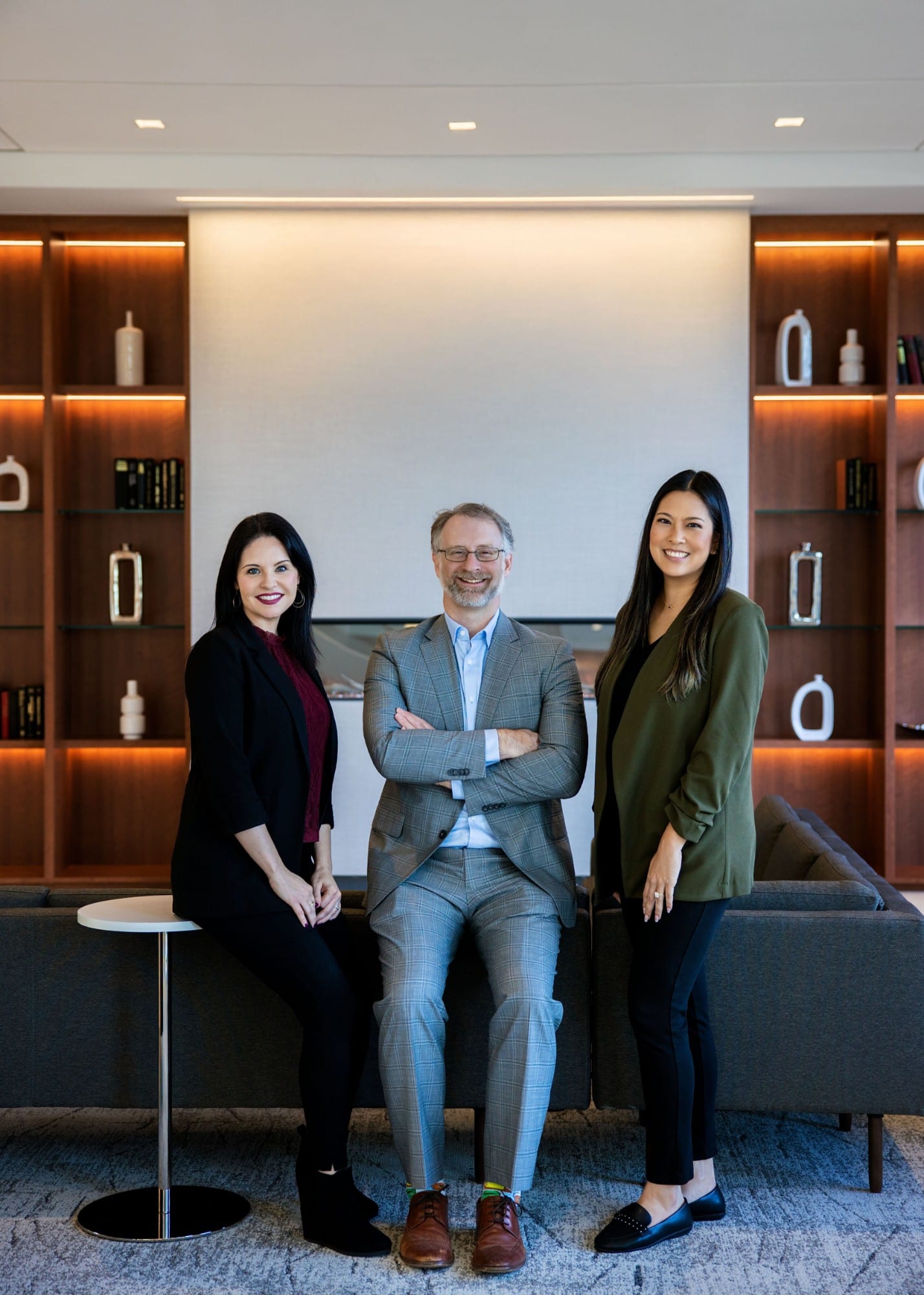 Three colleagues in a warmly lit lounge with wood shelving, two standing and one seated on a sofa arm
